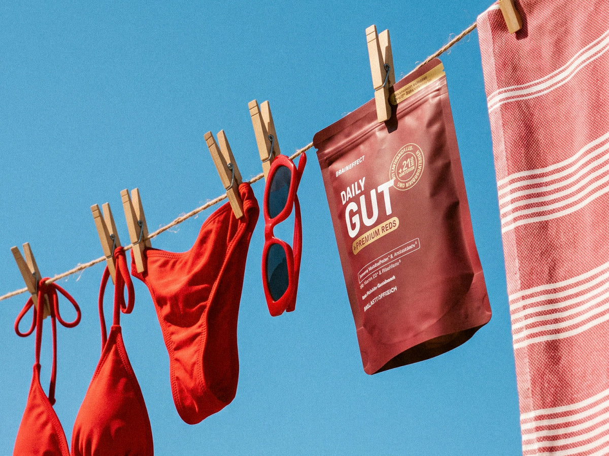 Eine BRAINEFFECT Daily Gut Premium Reds Beutel hängt an einer Wäscheleine im Freien mit der Aufschrift "BRAINEFFECT DAILY GUT PREMIUM REDS" neben roter Bikiniwäsche und einem Handtuch vor klarem blauen Himmel.