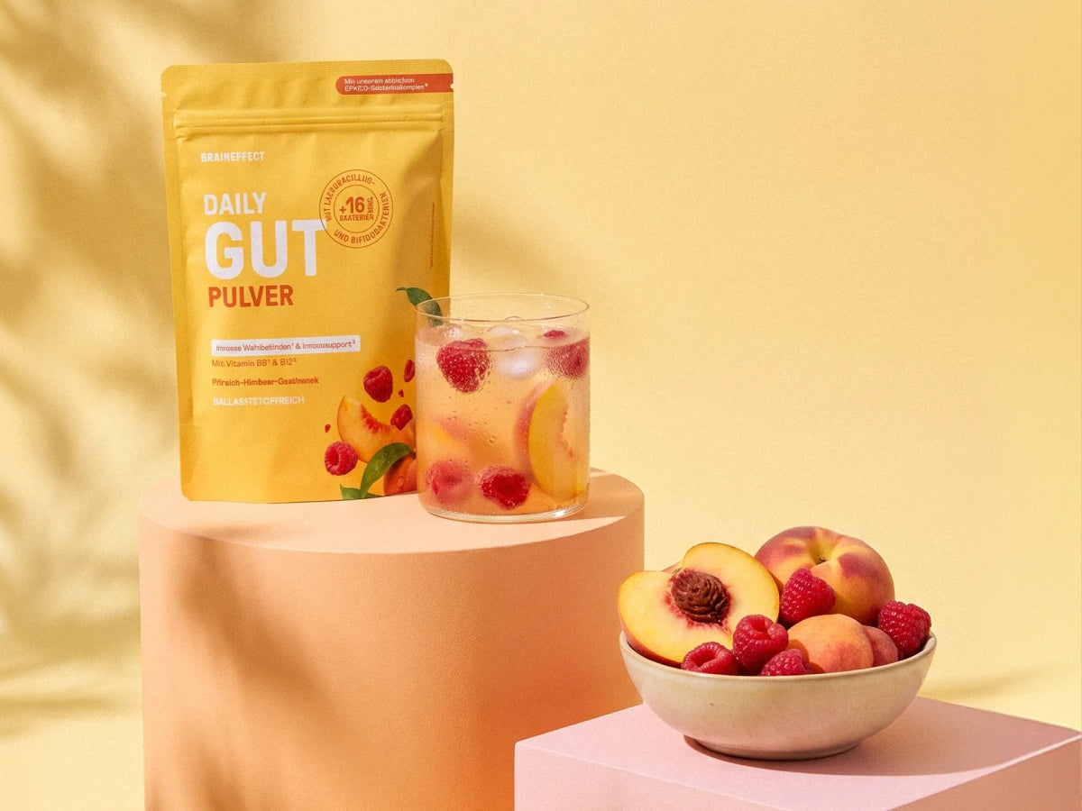 BRAINEFFECT DAILY GUT PULVER Verpackung neben einem Glas mit einem Pfirsich-Himbeer-Getränk, Eiswürfeln und frischen Früchten auf einem Podest