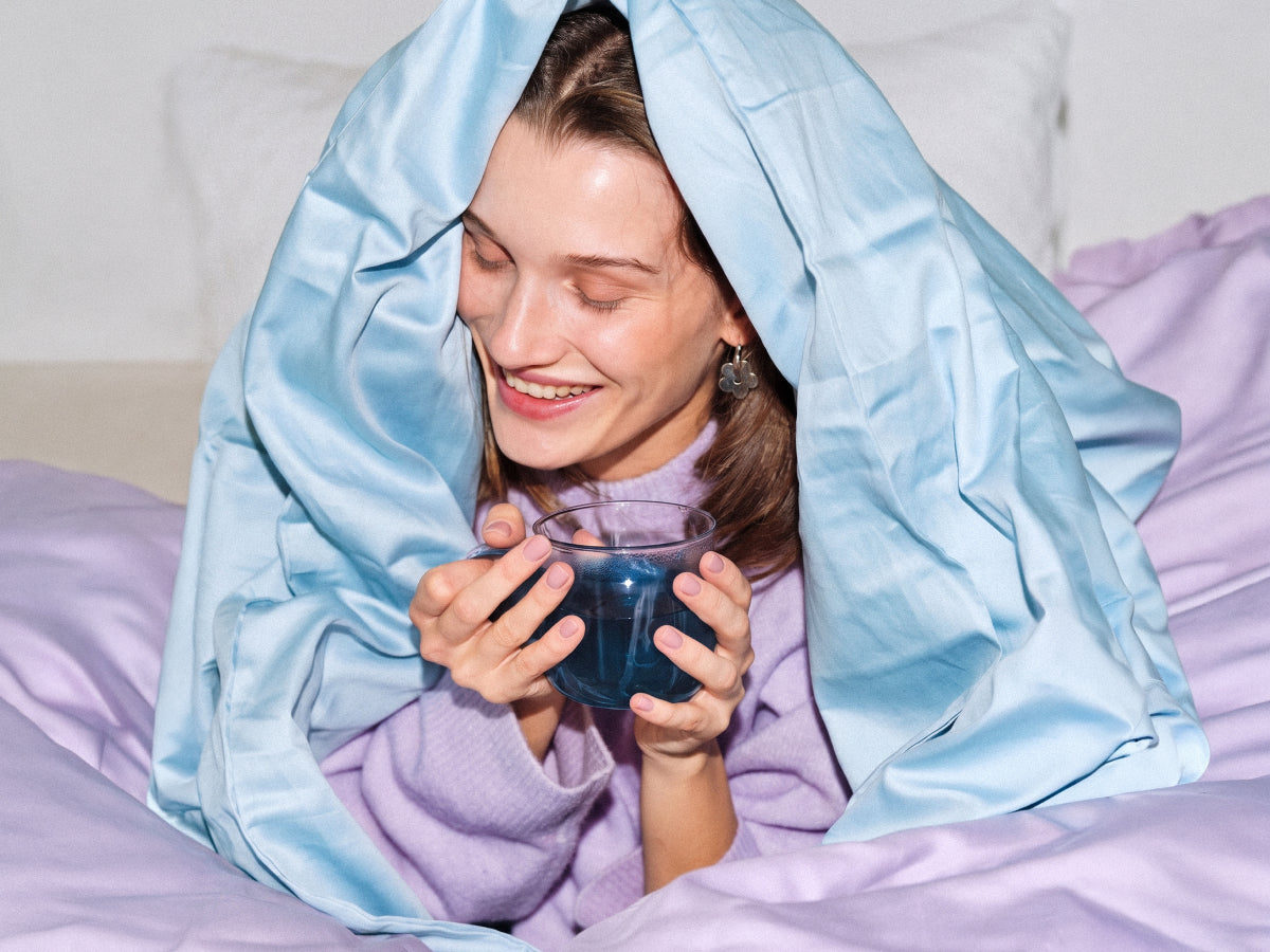 Eine lächelnde Frau sitzt im Bett, hat eine hellblaue Bettdecke über den Kopf gezogen und hält eine Tasse mit blauem Getränk in den Händen.
