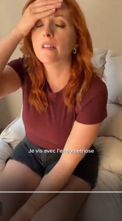 Charger la vidéo : Eine rothaarige Frau sitzt auf einem Sofa und hält sich mit schmerzverzerrtem Gesicht die Hand an die Stirn, mit dem Text "Je vis avec l'endométriose".