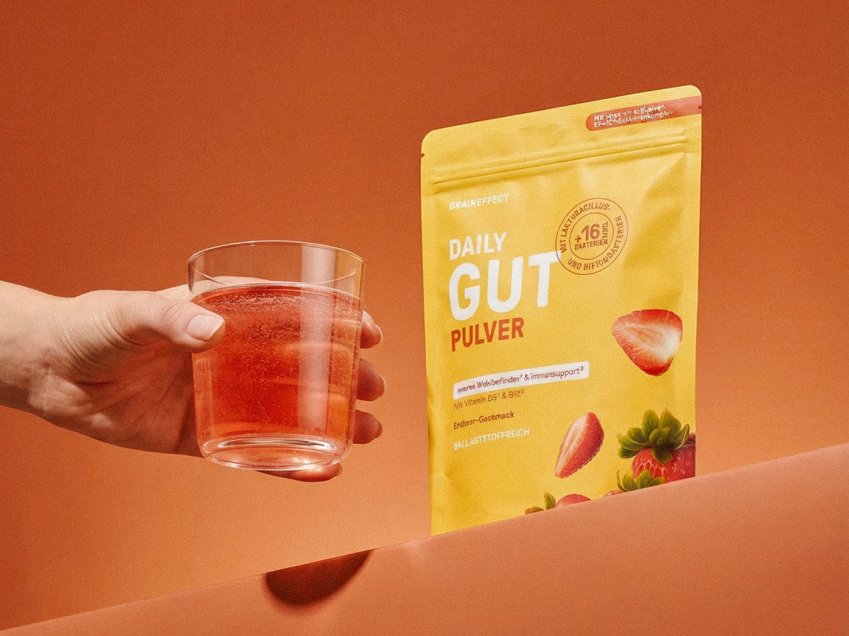 Eine Hand hält ein Glas mit rötlicher Flüssigkeit neben einem gelben BRAINEFFECT Beutel mit der Aufschrift "DAILY GUT PULVER", "inneres Wohlbefinden & Immundsupport", "mit Vitamin B6 & B12", "Erdbeer-Geschmack", "BALLASTSTOFFREICH", "BRAINEFFECT"