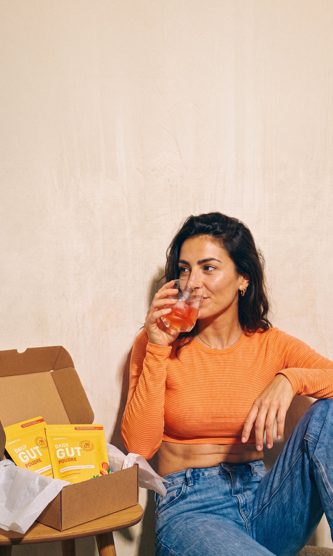 Eine Frau in orangefarbenem Oberteil trinkt aus einem Glas und blickt zur Seite, neben ihr auf einem Tisch stehen BRAINEFFECT "DAILY GUT POUDRE" Beutel in einem offenen Karton.