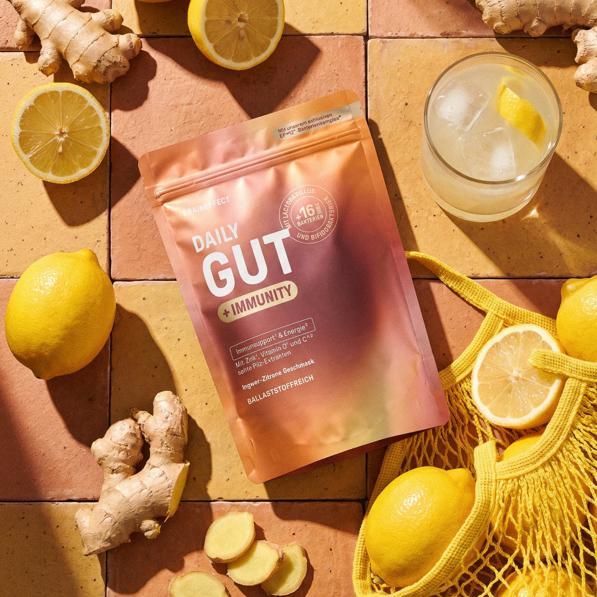 Produktbeutel BRAINEFFECT "DAILY GUT + IMMUNITY" umgeben von Zitronen, Ingwer und einem Glas auf hellen Fliesen mit dem Text "Mit unserem exklusiven EP>12*-Bakterienkomplex*", "MIT LACTOBACILLUS, +16 KULTUREN UND BIFIDOBAKTERIEN"