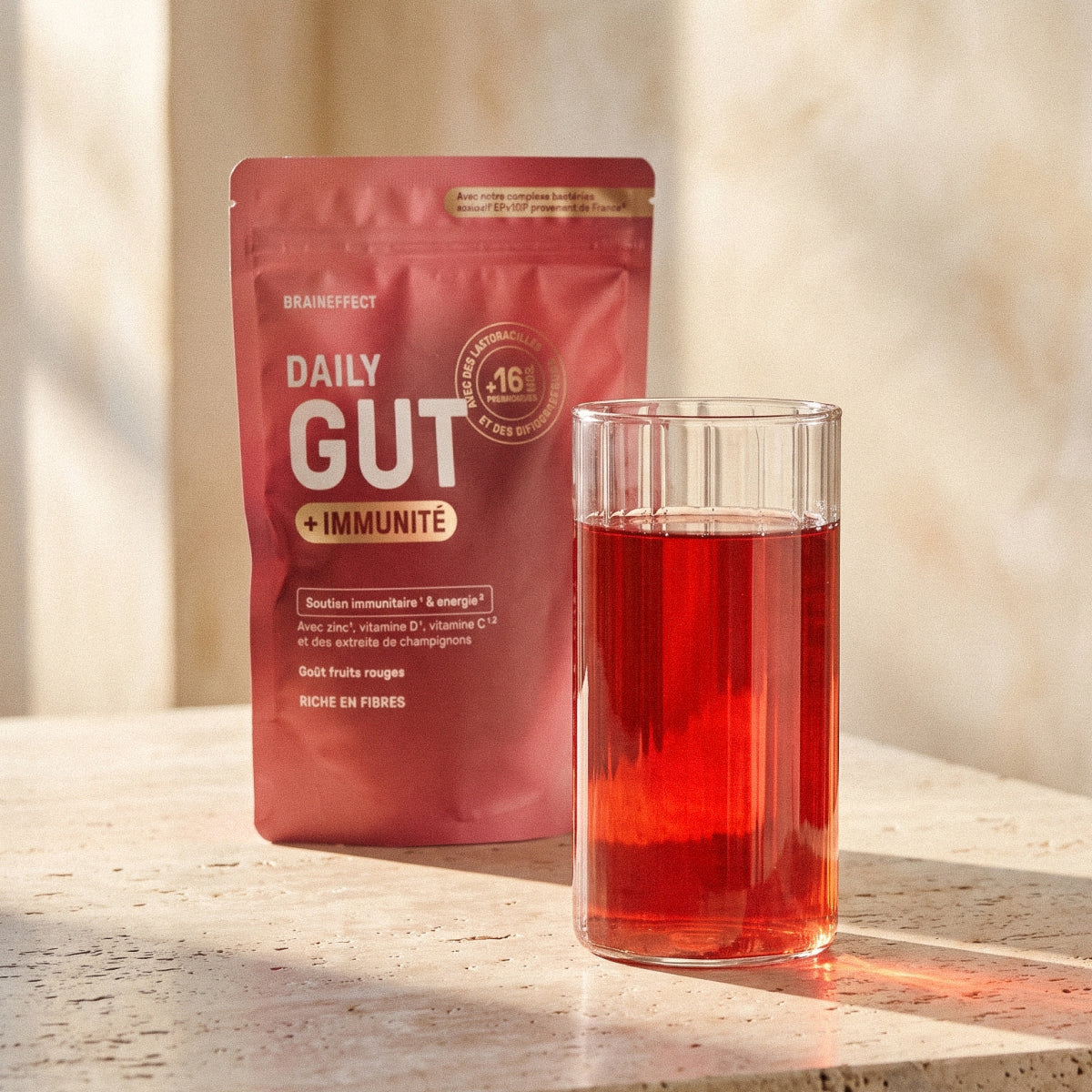 Sachet rouge BRAINEFFECT Daily Gut avec l'inscription "BRAINEFFECT DAILY GUT + IMMUNITÉ" à côté d'un verre de liquide rouge sur une table.