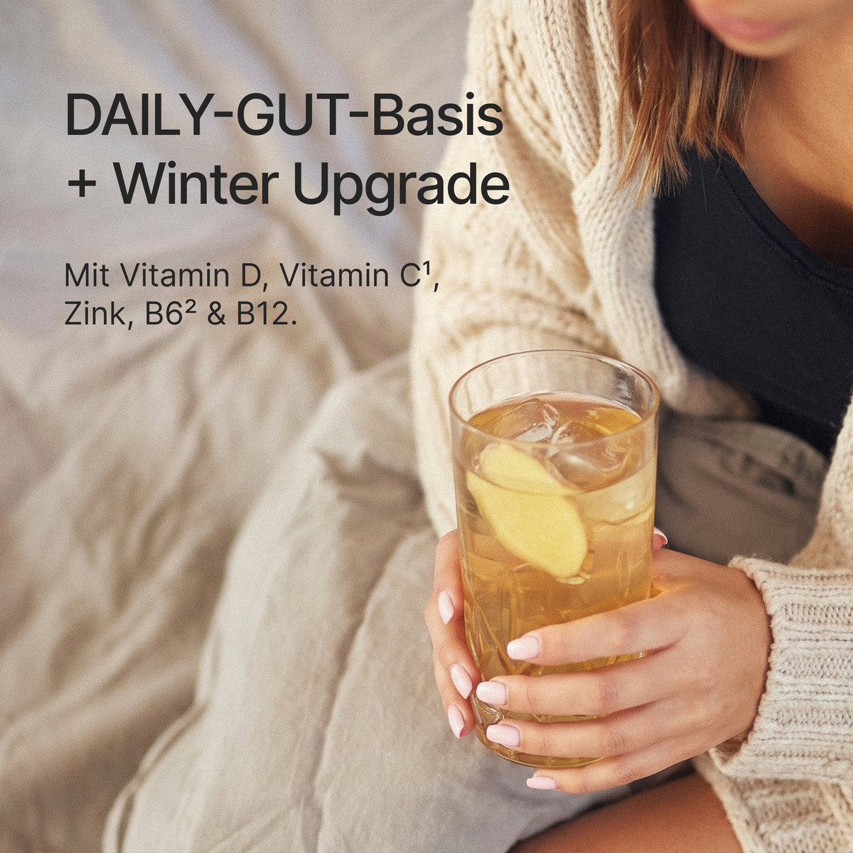 Femme en gilet tenant un verre de boisson claire avec une tranche de gingembre, accompagnée du texte "DAILY-GUT-Basis + Winter Upgrade" et "Mit Vitamin D, Vitamin C¹, Zink, B6² & B12."