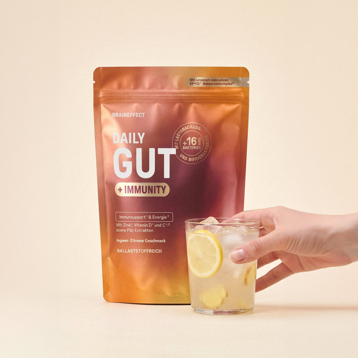 BRAINEFFECT Beutel mit Aufschrift "BRAINEFFECT DAILY GUT + IMMUNITY Mit unserem exklusiven EP×12®-Bakterienkomplex" vor beigem Hintergrund, daneben hält eine Hand ein Glas mit Eiswürfeln, Zitrone und Ingwer.