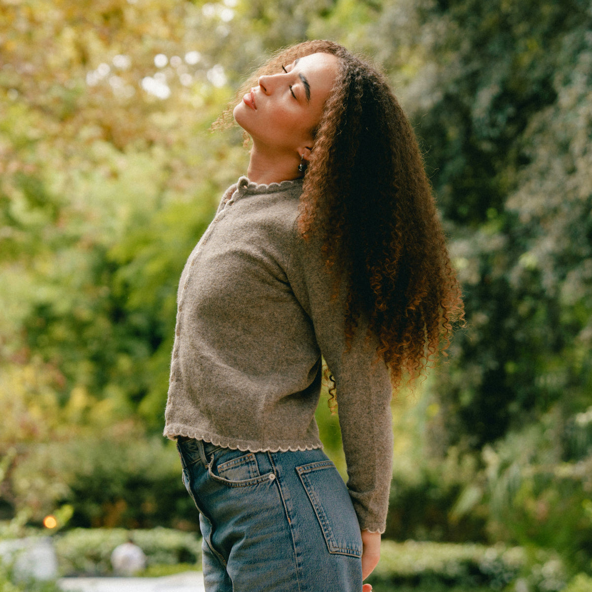 Jeune femme aux longs cheveux bouclés et aux yeux fermés se tient en plein air devant un fond vert avec la tête inclinée vers l'arrière
