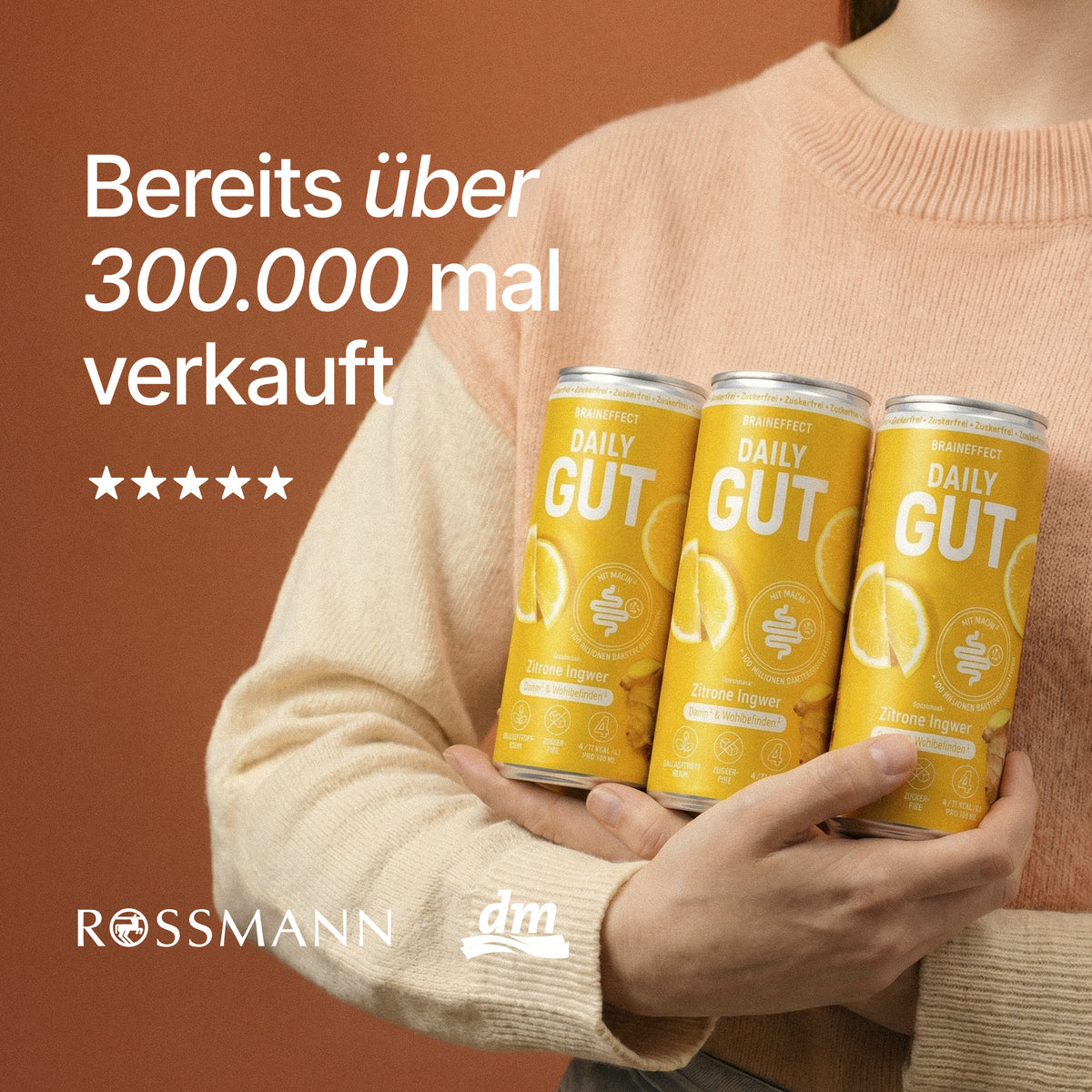 Person im beigen Pullover hält drei BRAINEFFECT Daily Gut Zitrone Ingwer Getränkedosen vor der Aufschrift "Bereits über 300.000 mal verkauft" sowie ROSSMANN und dm Logos