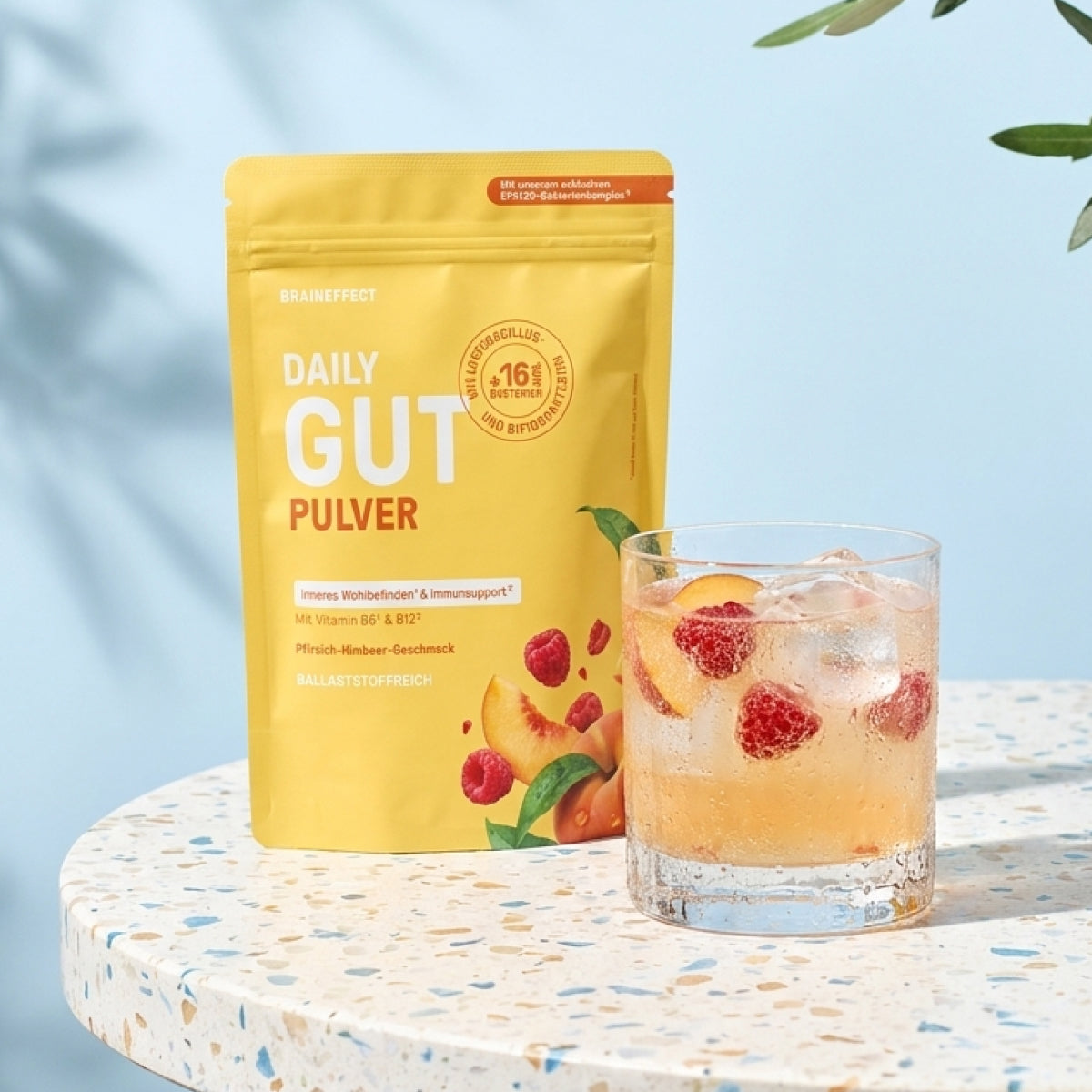 BRAINEFFECT DAILY GUT PULVER Standbeutel neben einem Glas Erfrischungsgetränk mit Eiswürfeln, Himbeeren und Pfirsich auf einem gemusterten Tisch im Sonnenlicht