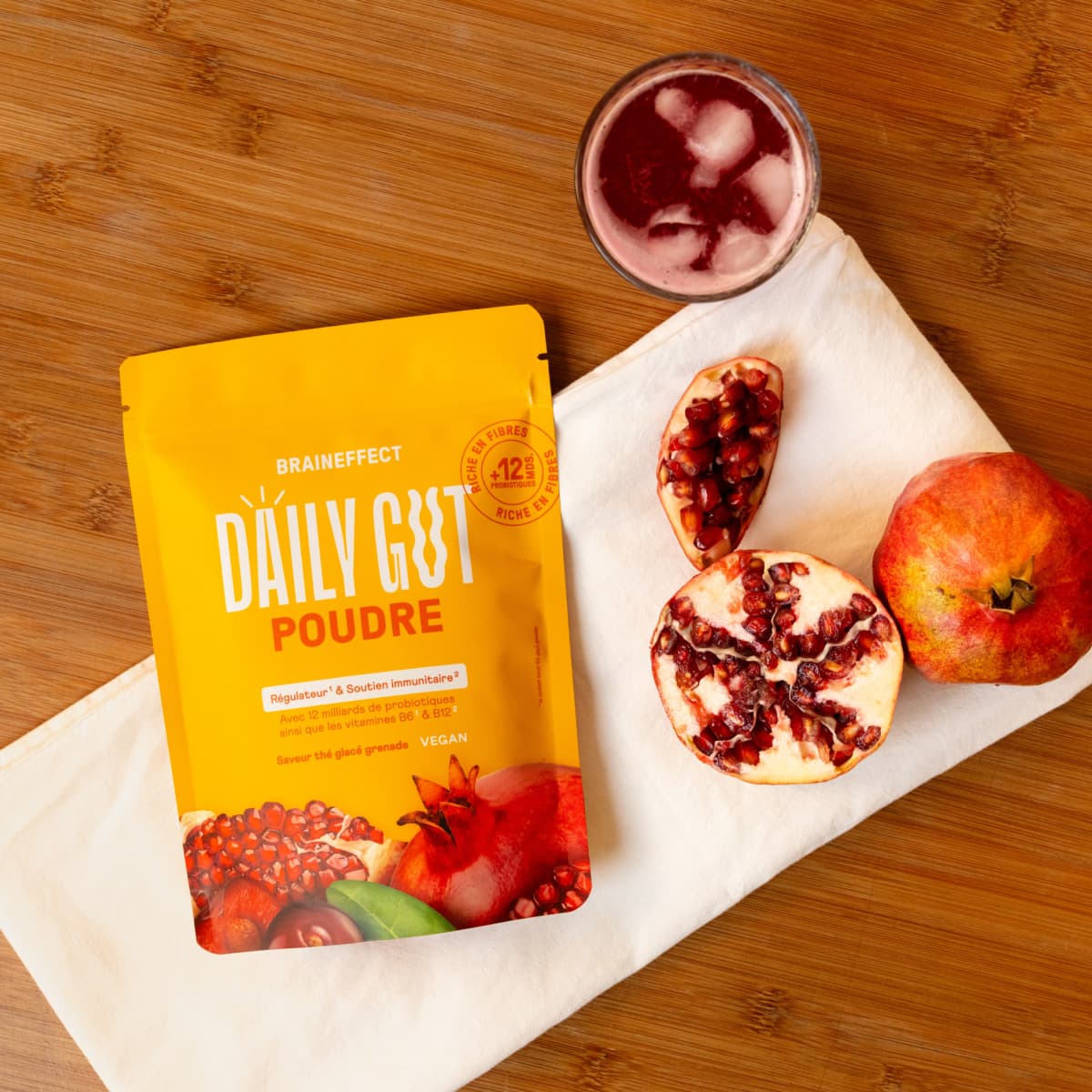 Eine gelbe Packung Daily Gut Puder von BRAINEFFECT liegt auf einem weißen Tuch, umgeben von Granatapfelhälften und einem Glas Pomegranate-Ice-Tea.