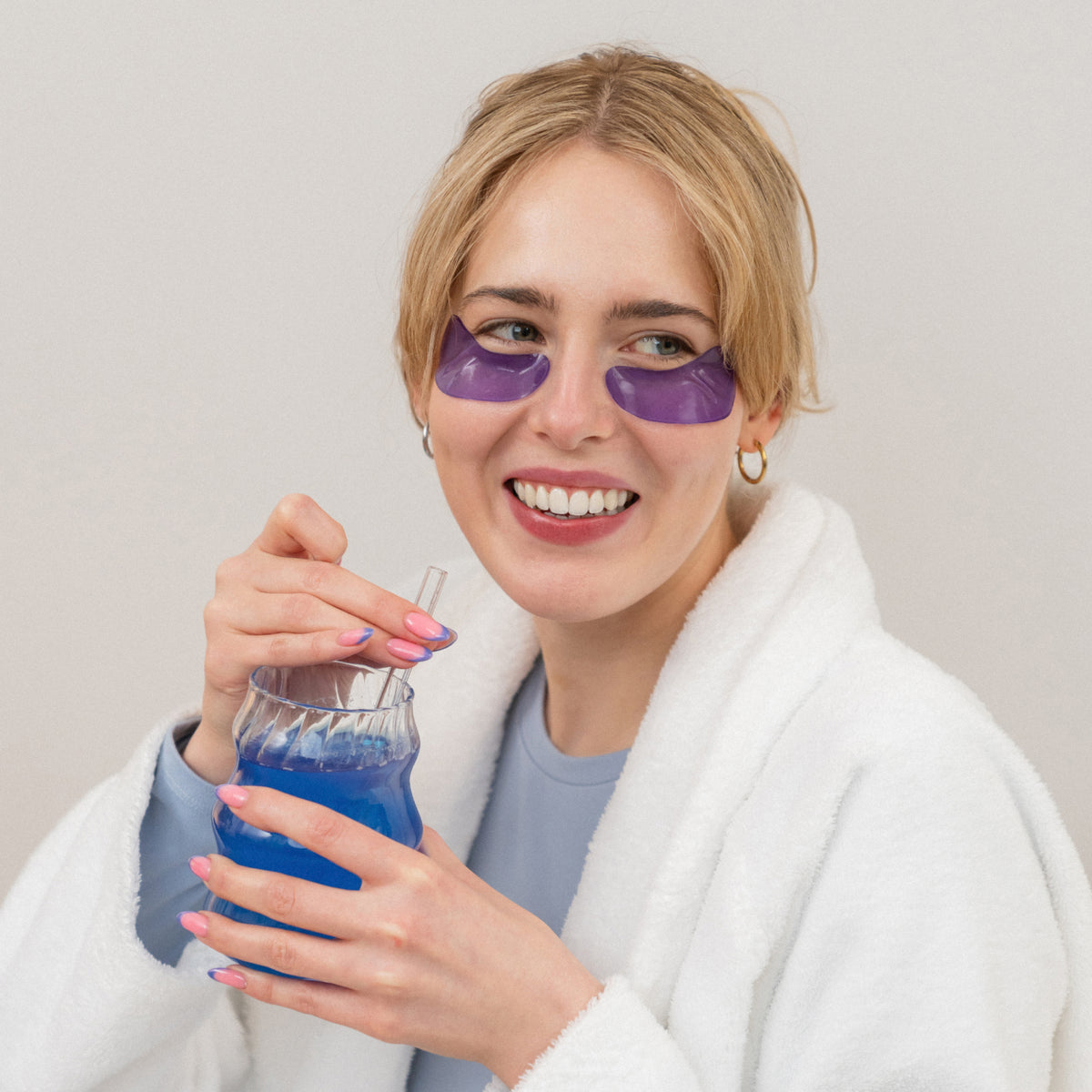 A blonde woman in a white bathrobe wears purple eye patches and drinks a blue beverage from a glass with a straw.