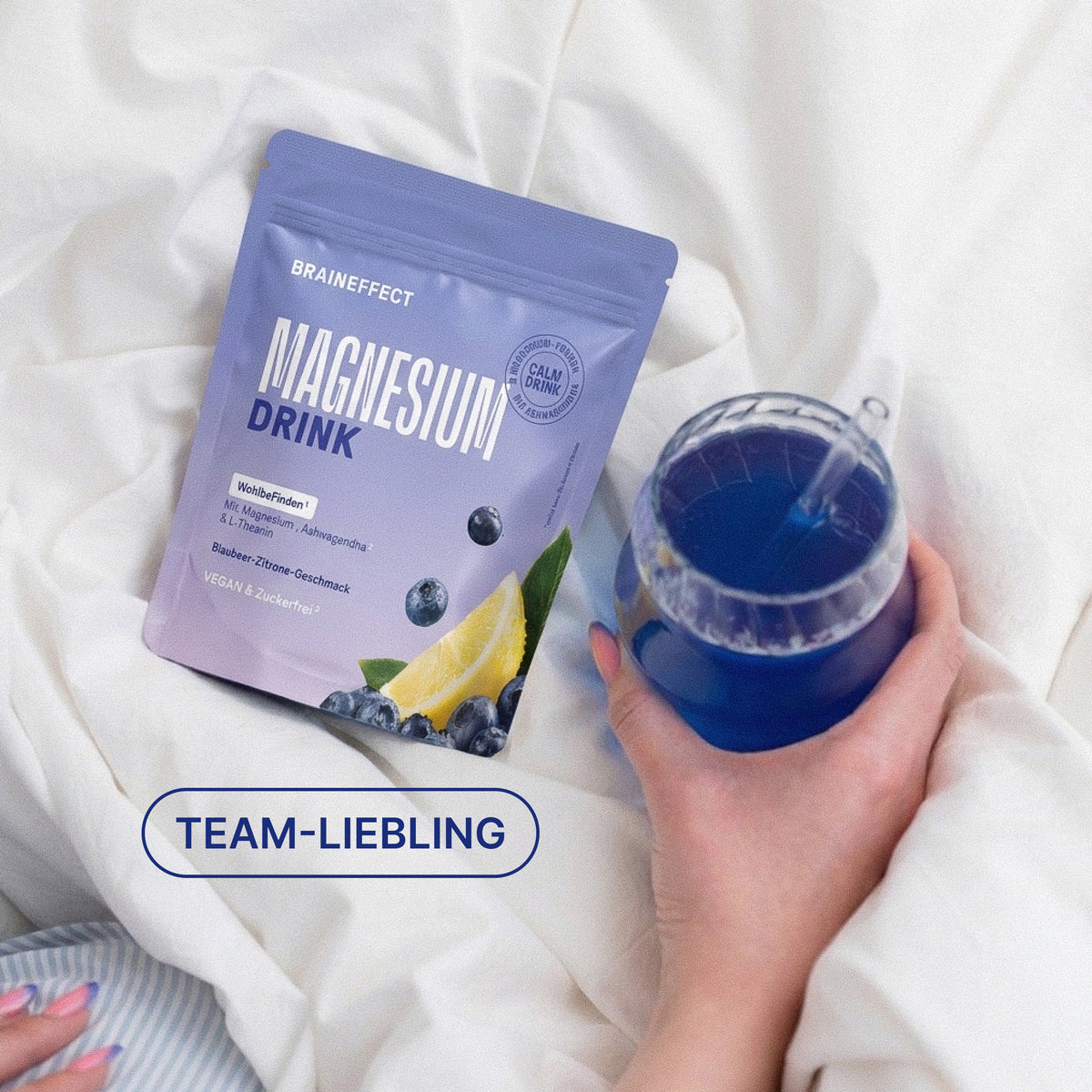 Das Badge "TEAM-LIEBLING" und die BRAINEFFECT-Packung "MAGNESIUM DRINK Mit Magnesium, Ashwagandha & L-Theanin Blaubeer-Zitrone-Geschmack VEGAN & Zuckerfrei" liegen auf weißer Bettwäsche neben einem blauen Getränk.
