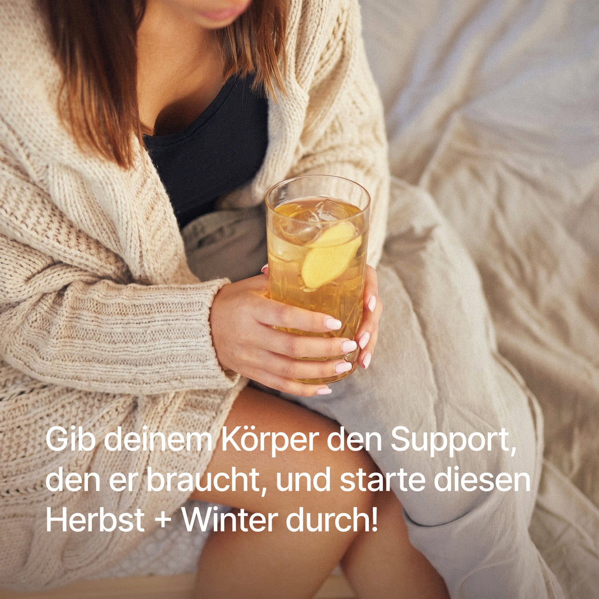 Frau in Strickjacke hält ein Glas mit einem Getränk, Ingwerscheibe und Eiswürfeln, mit dem Text "Gib deinem Körper den Support, den er braucht, und starte diesen Herbst + Winter durch!".