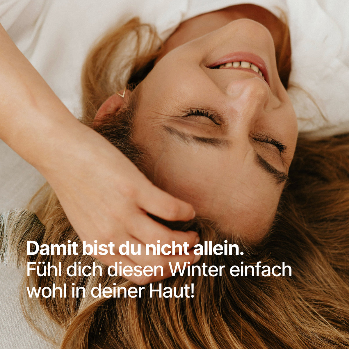 Frau liegt im Bett und lächelt mit dem Text "Damit bist du nicht allein. Fühl dich diesen Winter einfach wohl in deiner Haut!" von BRAINEFFECT
