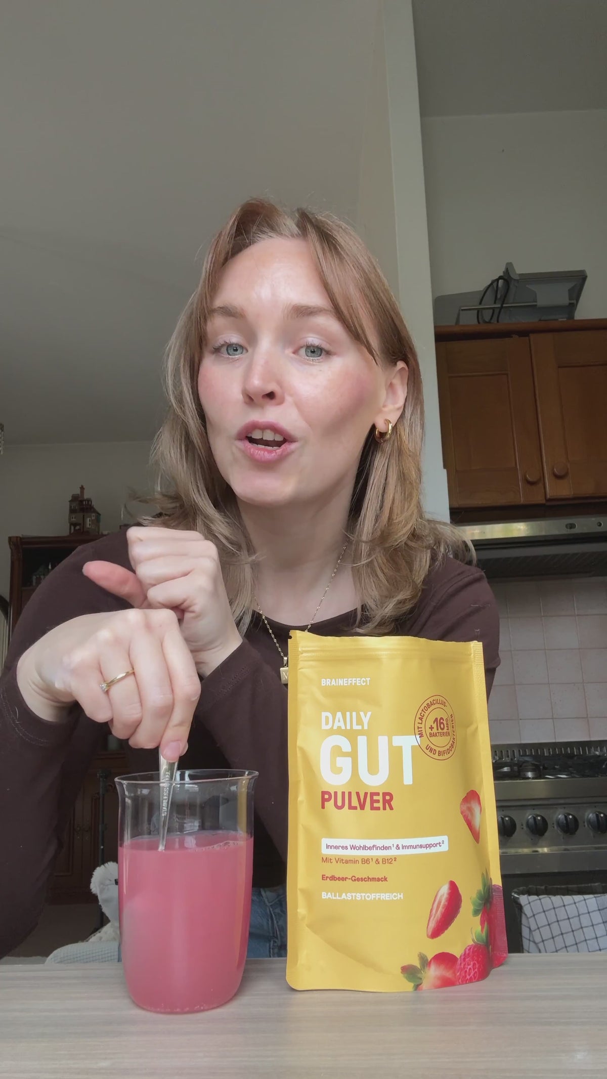 Video laden: Eine Frau rührt an einem Tisch in einem Glas mit einem pinkfarbenen Getränk, neben ihr steht ein gelber BRAINEFFECT Beutel mit der Aufschrift "DAILY GUT PULVER", "Inneres Wohlbefinden¹ &amp; Immunsupport²", "Mit Vitamin B6 &amp; B12³", "Erdbeer-Geschmack"