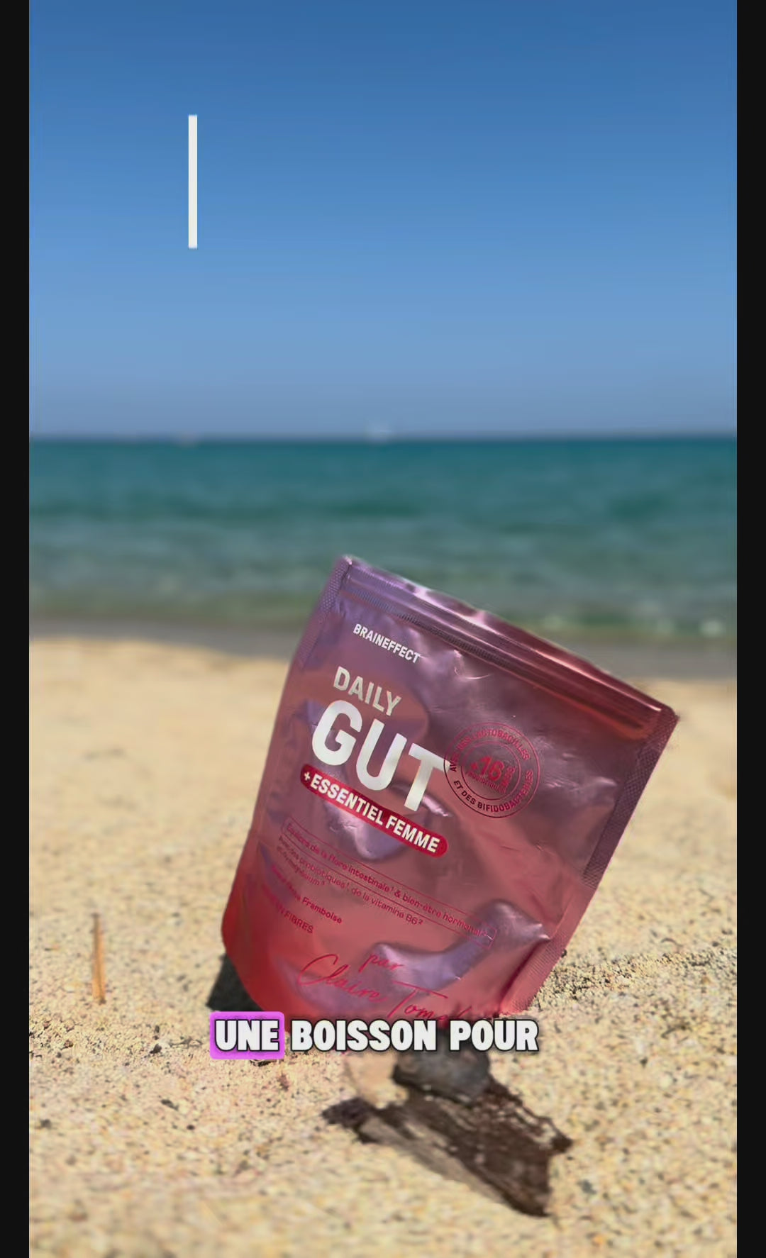 Charger la vidéo : Eine glänzende, rosafarbene BRAINEFFECT-Verpackung mit dem Text "DAILY GUT + ESSENTIEL FEMME" und "UNE BOISSON POUR" liegt im Sand am Strand vor dem Meer.