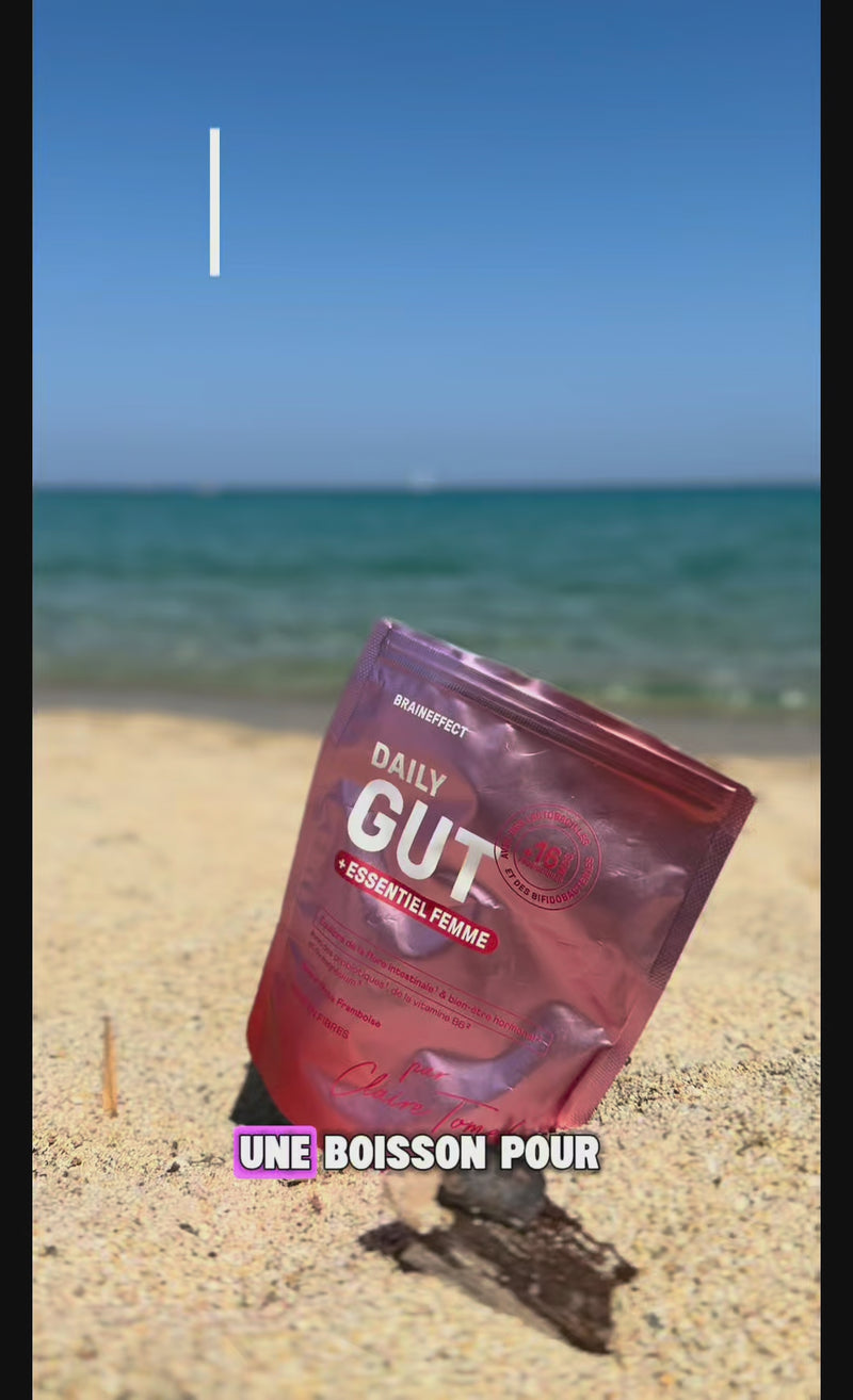 Eine glänzende, rosafarbene BRAINEFFECT-Verpackung mit dem Text "DAILY GUT + ESSENTIEL FEMME" und "UNE BOISSON POUR" liegt im Sand am Strand vor dem Meer.