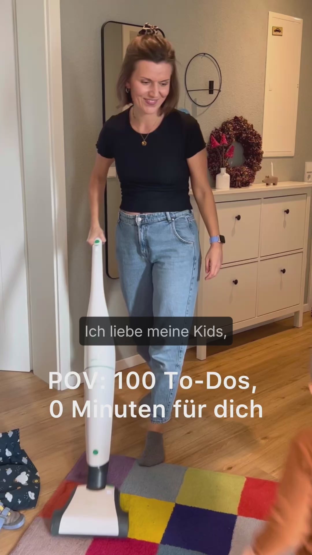 Video laden: Eine Frau in einem schwarzen T-Shirt und hellen Jeans saugt einen bunten Teppich, während die Aufschriften "Ich liebe meine Kids," und "POV: 100 To-Dos, 0 Minuten für dich" eingeblendet sind.