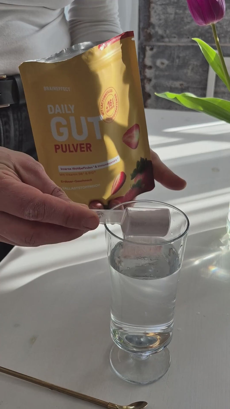 Eine Person hält einen BRAINEFFECT Beutel "DAILY GUT PULVER" und füllt Pulver mit einem Messlöffel in ein Glas Wasser.