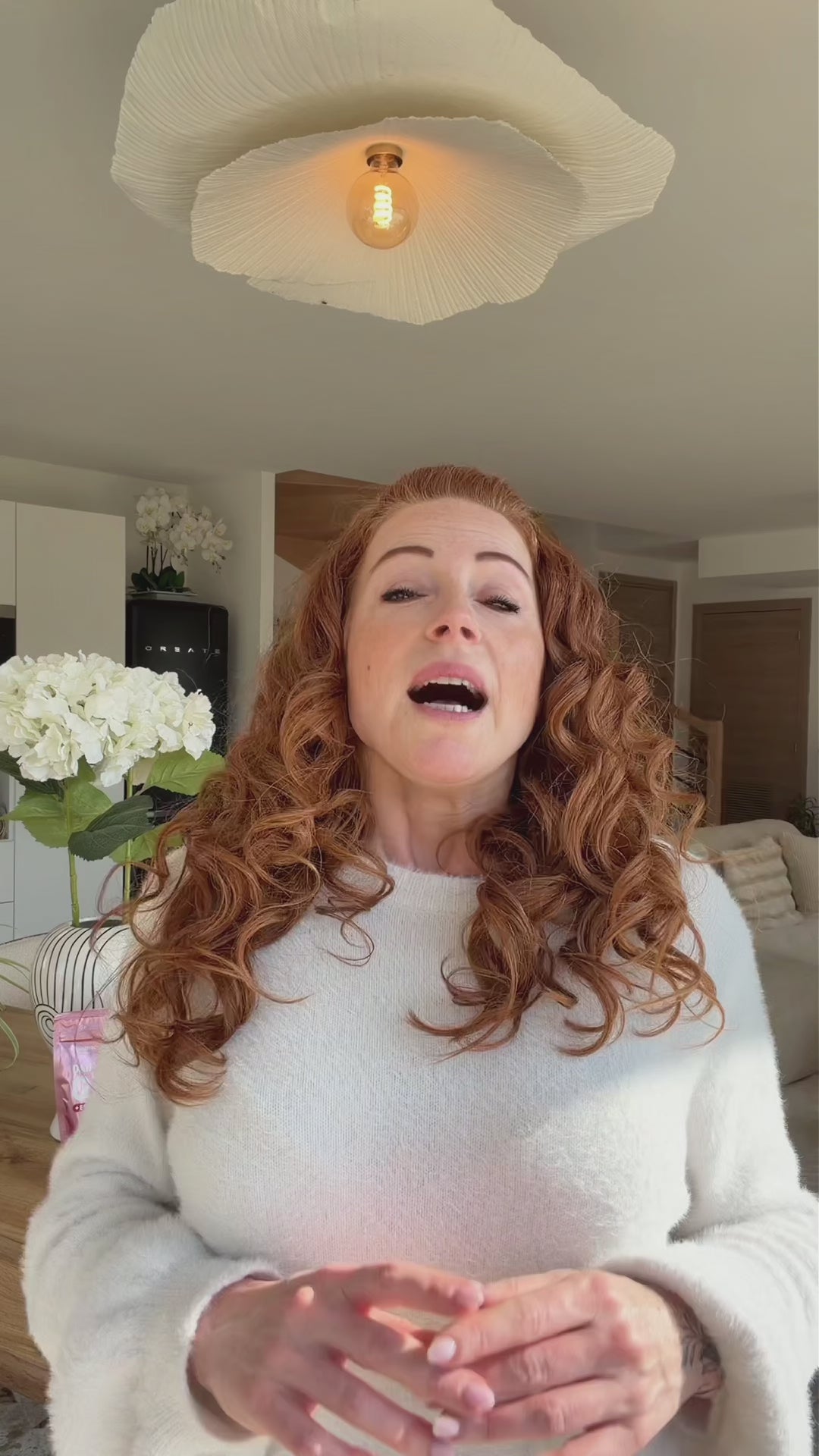 Charger la vidéo : Eine rothaarige Frau mit gelocktem Haar spricht in einem hellen Wohnzimmer mit weißer Deckenlampe.