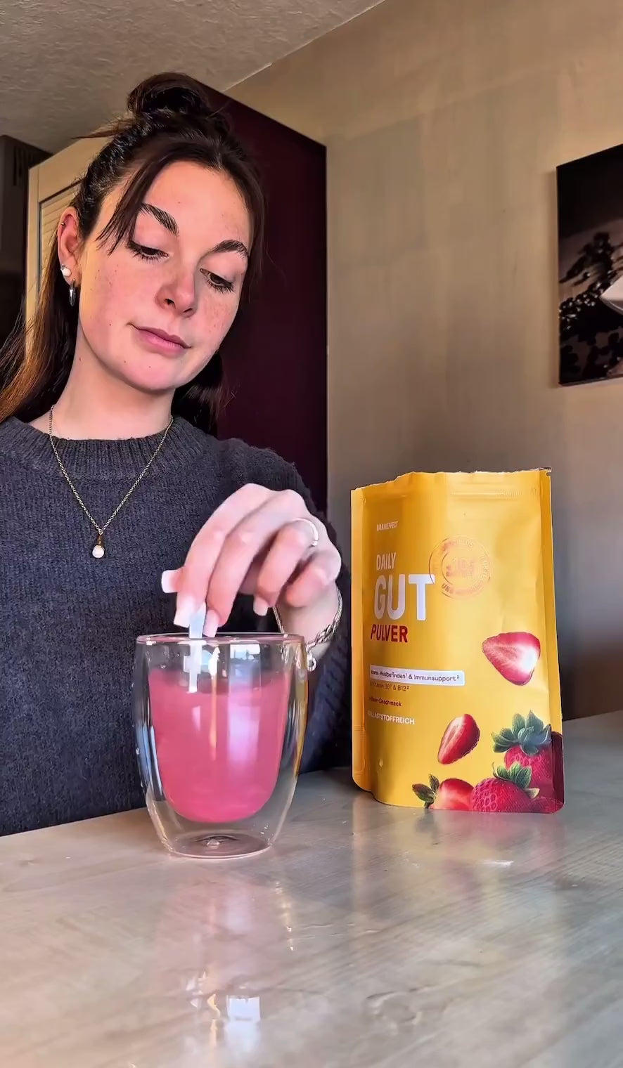 Video laden: Eine Frau in einem grauen Pullover rührt mit einem Löffel ein pinkes Getränk in einem Glas an, daneben steht ein gelber Beutel BRAINEFFECT "DAILY GUT PULVER".