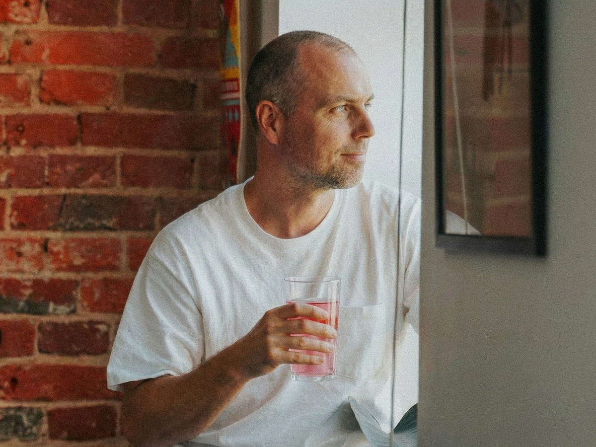 Ein Mann im weißen T-Shirt steht vor einer Ziegelwand, blickt aus einem Fenster und hält ein Glas mit pinkfarbenem BRAINEFFECT Getränk in der Hand