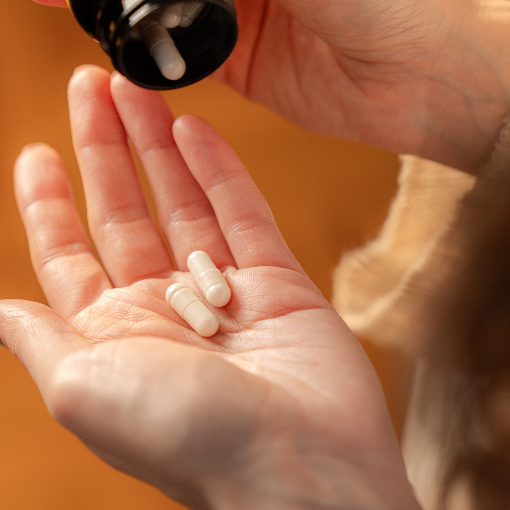 A hand holding two white capsules, with a bottle hovering above, representing daily wellness rituals.